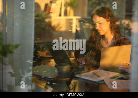 Concentrated telecommuter working at home using laptop and holding documents folder at sunset viewed through a window Stock Photo