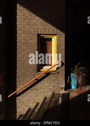A minimalist close-up of a brick wall featuring a yellow window frame and an orange handrail under strong geometric sunlight shadows. Stock Photo