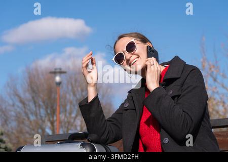Smiling woman wearing sunglasses and a black coat, talking on a mobile phone while resting with her suitcase on a bench, communicating with loved ones Stock Photo