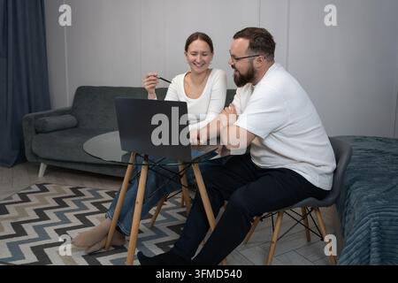 Young adult couple collaborating and sharing ideas, analyzing financial papers and using a laptop while sitting at a glass table in their modern home Stock Photo