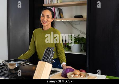 African American woman standing in kitchen in olive sweater, chopping purple cabbage by knife block Stock Photo