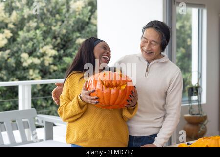 Senior Asian man and woman holding carved jack-o'-lantern on covered deck, standing in sweaters Stock Photo