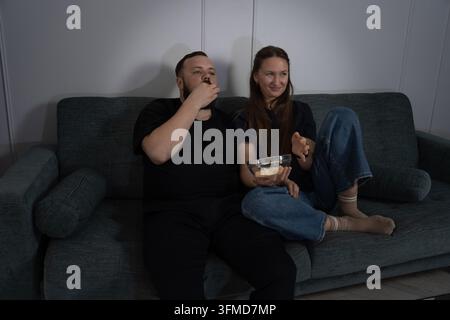 Couple sitting on couch in dimly lit room, sharing snacks and immersed in watching TV, while woman smiling and discussing content with her partner Stock Photo