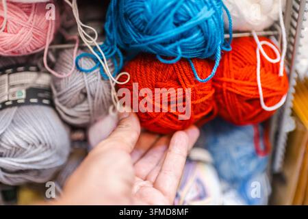 A hand touches a bright red ball of yarn among colorful yarn on a store shelf. Stock Photo