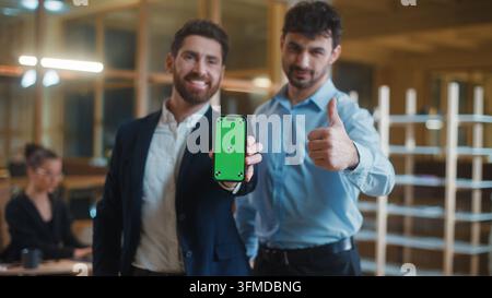 Two Caucasian business men smiling happy showing mobile phone with green screen demonstrate thumb up like gesture cheerful positive businessmen male Stock Photo