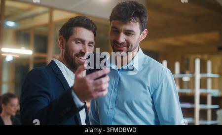 Two Caucasian business men looking in mobile phone screen watching online content smiling laughing in office. Cheerful businessmen male colleagues Stock Photo
