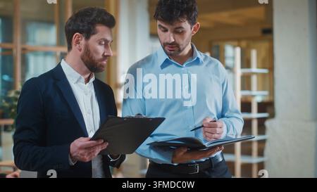 Two Caucasian business men comparing data with documents talking discussing stats in office. Focused businessmen executives colleagues talk discussing Stock Photo