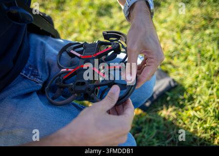 Pilot's hands holding a small FPV quadcopter, checking propeller guards and blades against blurred green grass, preparing equipment for outdoor flight Stock Photo