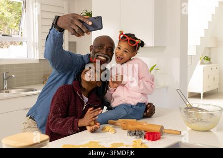 African family senior man and kids taking Christmas cookie selfie kitchen counter with rolling pin Stock Photo