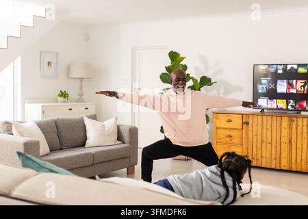 Mature man practicing yoga in living room wearing pink hoodie while child on sofa watching TV Stock Photo
