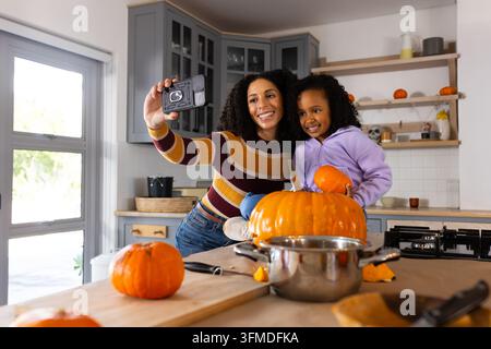 African American mom and youth daughter taking selfie in kitchen with halloween pumpkin, camera Stock Photo
