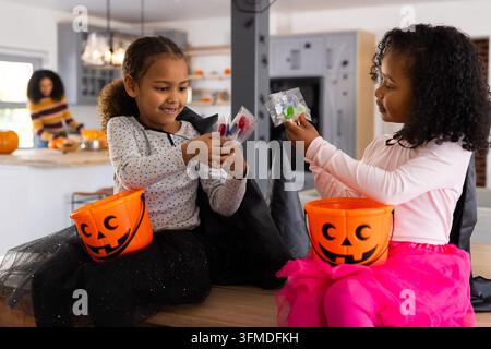 African American female family, two caped kids sharing halloween candies with pumpkins in kitchen Stock Photo