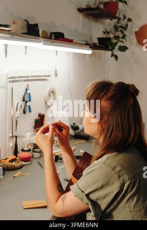 Female leatherworker prepares thread while seated at a craft bench with leather in a stitching clamp. Skilled handmade production and creative work co Stock Photo