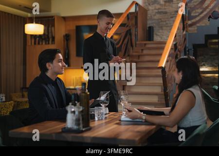 Young male waiter taking orders from a couple, Caucasian man and Asian woman sitting at a luxury restaurant table, side view. Stock Photo