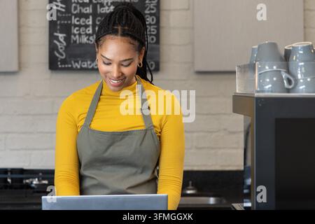 African American woman in gray apron smiling while typing on laptop at counter with coffee machine. Barista, cafe, espresso, chalkboard, brick, mug, warmth Stock Photo