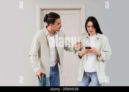 Relationship conflict and communication breakdown, man with power tool interrupting woman using smartphone during home renovation project in living ro Stock Photo