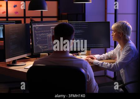 Software development and coding collaboration, programmers working late on project deadline in dark office with multiple computer monitors and code Stock Photo
