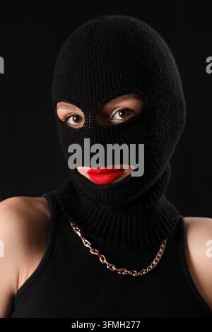Cybersecurity and digital crime, woman wearing black balaclava mask with red lipstick and gold chain, anonymous hacker or protest in dark studio Stock Photo