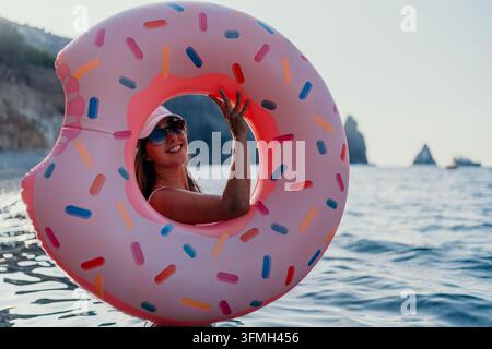 Woman donut ocean. Happy woman holding pink inflatable donut float in shimmering ocean during summer. Stock Photo