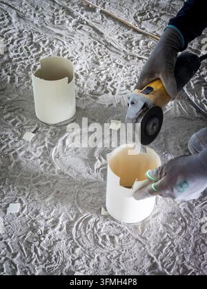 Construction worker wearing protective gloves uses an angle grinder to cut white PVC conduit sections on a dusty unfinished floor. Electrical cables, Stock Photo