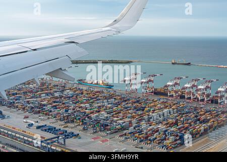 Aerial view from an airplane window over a busy commercial seaport in Spain showing cargo ships, cranes and rows of shipping containers during a fligh Stock Photo