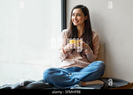 Happy latin woman enjoying coffee at cafe, sitting near window and looking into it, empty space. Smiling dreamy lady holding mug with hot beverage. Le Stock Photo