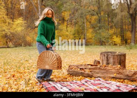 Woman carrying wicker basket near picnic blanket in autumn park Stock Photo