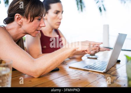 two focused young women doing remote work on laptop at outdoor terrace as digital nomad lifestyle in an outdoor cafe bar Stock Photo