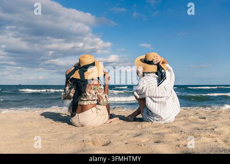 Rear view of a senior woman and a young woman of different ethnic backgrounds sitting on the beach, holding their straw hats against the wind as they Stock Photo