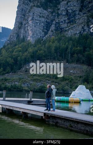 A man and a woman are standing on a lake dock and looking at the mountains. Stock Photo