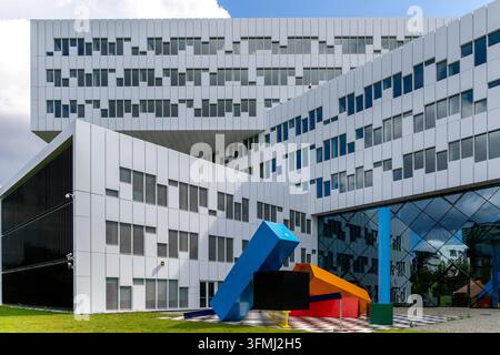 Modern white office building with colorful geometric sculpture in a sunny courtyard, blue sky, green lawn and black-and-white checkerboard pavement. Stock Photo