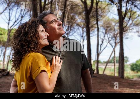 A woman rests her hand on her partner's chest as they look upward together in a pine forest, suggesting affection, trust and a shared outlook. Stock Photo