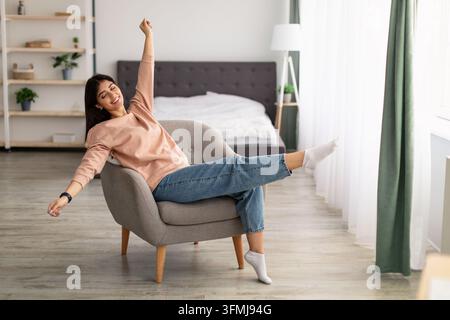 People, Leisure and Relaxation concept. Indoor shot of happy beautiful woman freelancer reclining on chair, raising arms in the air while stretching b Stock Photo