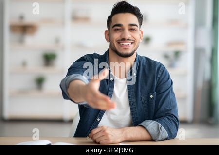 Positive arab man sitting at workdesk, giving hand and smiling at camera, office interior, copy space. Happy young middle-eastern guy sitting at table Stock Photo