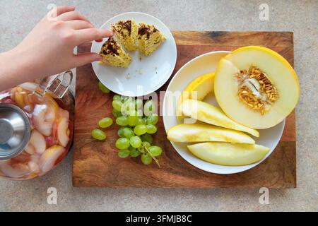 a woman's hand with a piece of homemade cake, melon slices, grapes and stewed fruit in glass teapot on a wooden background, concept of fresh fruits an Stock Photo
