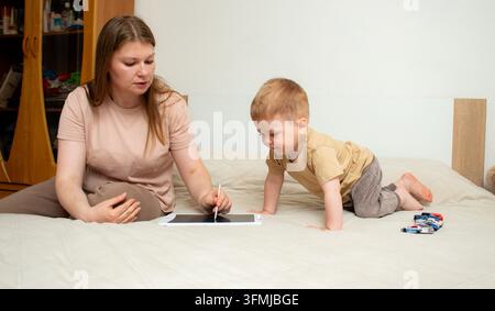 Toddler drawing on tablet using stylus, mom guiding his hand on tablet screen. Early creative development through shared activity. Parent child game s Stock Photo