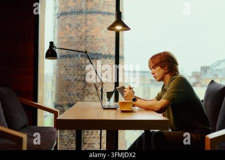Man Writing in Notebook While Working on Laptop in Cafe Stock Photo