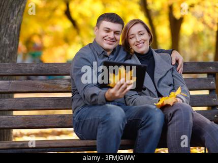 a woman and a man are sitting on a bench in an autumn park, they have a pad and a bouquet of yellow maple leaves, a bright sunny day Stock Photo
