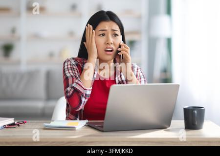 Oh No. Shocked Asian Female Freelancer Talking On Cellphone At Home Office, Frustrated Korean Woman Sitting At Desk With Laptop, Emotionally Reacting Stock Photo