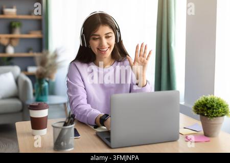Online Meeting Concept. Young smiling woman making video call on laptop at home, positive female in wireless headset waving at computer web camera, en Stock Photo