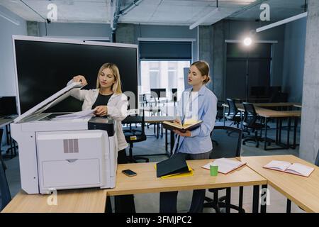 Two businesswomen collaborating and reviewing documents in a modern office, standing by a multifunction printer Stock Photo