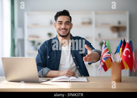 Cheerful arab guy foreign language teacher sitting at workdesk with laptop and bunch of international flags and giving hand. Smiling migrating service Stock Photo