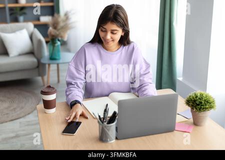 Business And Telecommuting Concept. High angle view of smiling young woman using her smartphone and modern laptop computer, reading text message, sitt Stock Photo