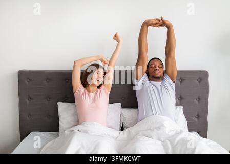 Multinational couple stretching on bed after waking up, feeling sleepy at home. Diverse young family being lazy on weekend morning, relaxing on Sunday Stock Photo