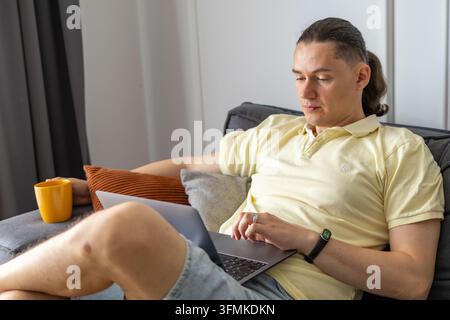 Thoughtful businessman working on laptop from home sofa while taking tea break from digital tasks. Concept of remote work, work life balance, producti Stock Photo