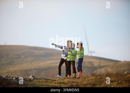 Three engineers in safety vests and helmets consult plans while surveying a wind farm on rolling hills, collaborating on renewable energy installation Stock Photo
