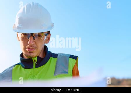 Focused construction worker in a hard hat and safety glasses studies blueprints while wearing a high-visibility vest on an outdoor job site, conveying Stock Photo