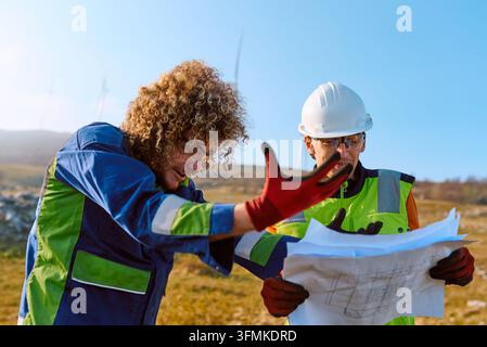 Two field engineers in high-visibility clothing and hard hats review technical plans outdoors at a wind farm, collaborating on installation details an Stock Photo
