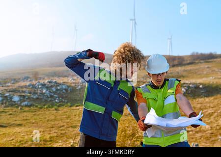 Two engineers in high-visibility vests and hard hats study blueprints on a hillside wind farm, discussing installation, inspection and maintenance of Stock Photo