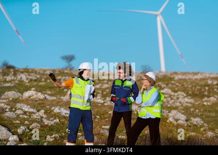 Three engineers in hard hats and high-visibility vests inspect a wind farm, reviewing plans and discussing site assessment, safety and project details Stock Photo